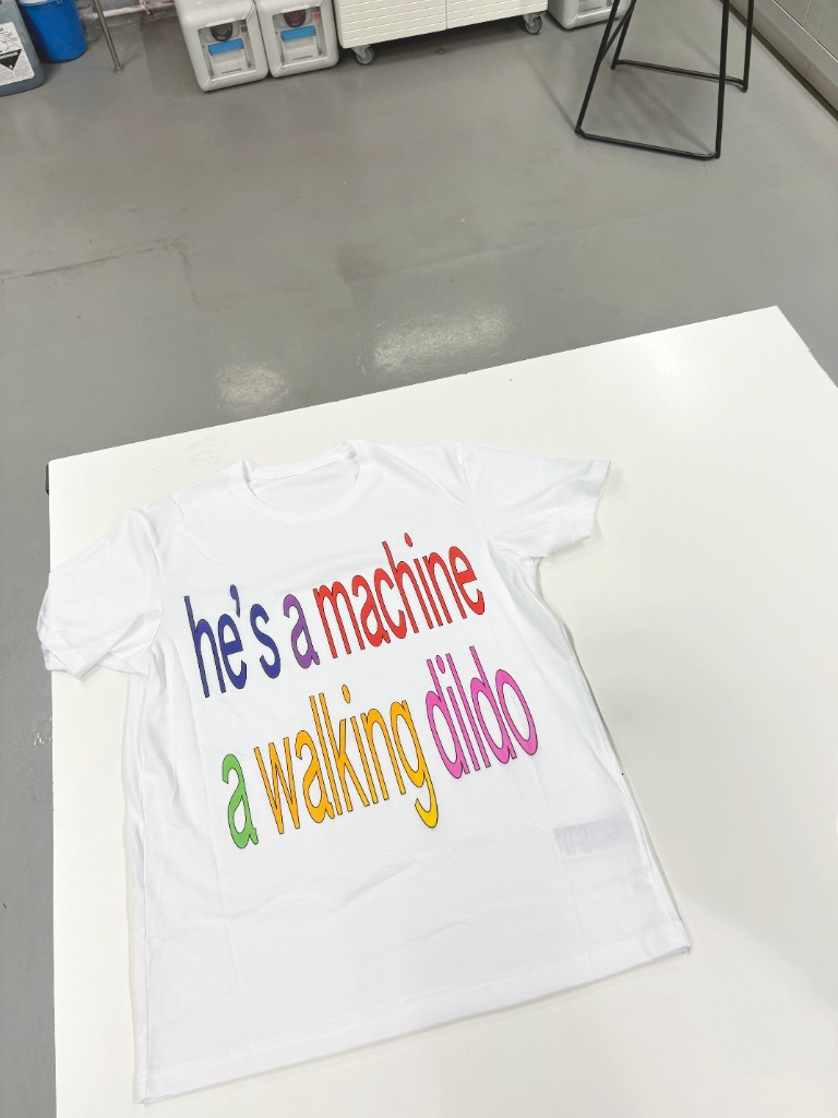 White t-shirt laid flat on a table, multicolour lettering: he's a machine, a walking dildo